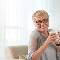 Portrait of happy mature woman in eyeglasses resting on sofa with cup of coffee and smiling at camera at home